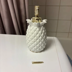 Vit ananasformad parfymflaska - Säljer en parfymflaska i form av en ananas. Flaskan är vit med guldfärgad pump och detaljer. Tillverkad i keramik eller porslin och har ett dekorativt utseende som sticker ut i badrummet eller på sminkbordet.