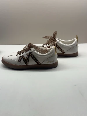 Steve Madden vita sneakers leoparddetaljer - Snygga vita sneakers från Steve Madden med leopardmönstrade detaljer och matchande skosnören i mesh. Skorna har en låg siluett, beige sula och gulddetalj på hälen. Perfekta för dig som vill sticka ut med coola detaljer och trendig look. Sko box kommer med. Skulle säga att dom passar både 37 och 38
