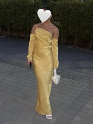 Guldig långklänning i satin - Snygg guldig långklänning i satin med one shoulder-design och långa ärmar. Klänningen har en elegant drapering över bysten och en figurnära passform som ger en lyxig känsla. Perfekt för speciella tillfällen när du vill sticka ut.