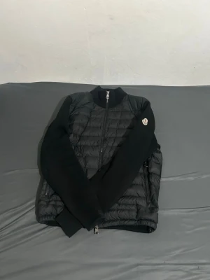 Svart cardiganjacka från Moncler - Svart quiltad cardiganjacka från Moncler med stickade ärmar och rygg. Jackan har dragkedja framtill, två sidofickor med dragkedja och Moncler-logga på ena ärmen. Perfekt för lager-på-lager och stilren look.