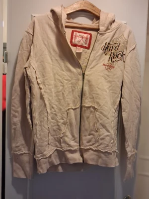 Beige Hard Rock zip hoodie - Säljer en beige hoodie från Hard Rock med dragkedja och tryck både fram och bak. Hoodien har huva, två fickor framtill och coola detaljer med slitna kanter. Materialet känns mjukt och bekvämt, perfekt för en avslappnad rockig stil.står xl men passar både M och L