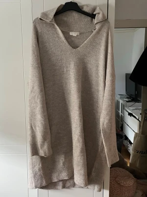 Beige stickad klänning  - Mysig beige stickad klänning och djup V-ringning. Tröjan har långa ärmar och ribbade muddar vid ärmslut och nederkant. Perfekt för lager på lager och chill stil. 