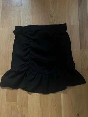 Svart volangkjol med rynk - Kort svart kjol med volangkant och rynkade detaljer framtill. Kjolen har en feminin och trendig look, perfekt att matcha med en enkel topp eller stickad tröja. Tillverkad i mjukt tyg för skön passform.