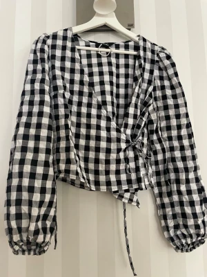 Svartvit rutig omlottblus - Trendig svartvit rutig omlottblus med långa puffärmar och knytband i midjan. Blusen har en djup v-ringning och är tillverkad i ett lätt, vävt material som ger en luftig känsla. Perfekt för att skapa en cool och avslappnad look.