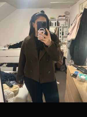 Kort brun dubbelknäppt kappa - Säljer en kort brun kappa med breda slag och dubbelknäppning. Kappan har stora knappar och uppvikta ärmslut med knappdetalj. Perfekt för dig som vill ha en stilren och trendig look. Materialet känns tjockt och varmt.