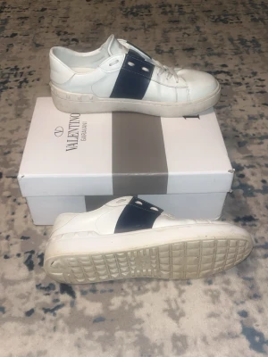 Valentino vita sneakers med blå detalj - Snygga vita sneakers från Valentino med bred mörkblå läderdetalj över vristen och vita snören. Klassisk låg modell med diskreta nitar på sulan och stilren design. Perfekta för dig som gillar exklusiva och trendiga skor.