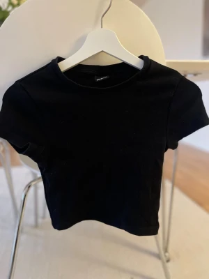 Svart croppad t-shirt från Gina Tricot - En svart croppad t-shirt från Gina Tricot med rund halsringning och korta ärmar. Toppen är enkel och stilren, perfekt att matcha med jeans eller kjol. Materialet känns mjukt och stretchigt för en bekväm passform.