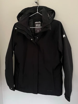 Svart vindjacka från Helly Hansen - Ny Helly Hansen Helly Tech regnjacka för dam i storlek S.  Jackan är svart och tillverkad i polyester med Helly Tech Protection-material, vilket ger ett vatten- och vindtätt men ventilerande yttertyg. Modellen har huva, hel dragkedja framtill med skyddande vindslå samt långa ärmar. Yttertyget består av polyester med beläggning, och fodret är också i polyester.  Passar som regnjacka och skaljacka vid vardagsbruk och utomhusaktiviteter i blött och blåsigt väder.