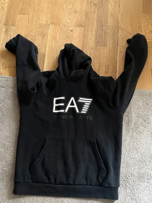 Svart EA7 hoodie från Emporio Armani - Snygg svart hoodie från Emporio Armani med stor EA7-logga i vitt på bröstet. Klassisk känguruficka framtill och huva med dragsko. Perfekt för en avslappnad och trendig stil. Tillverkad i mjukt material som känns skönt mot huden.