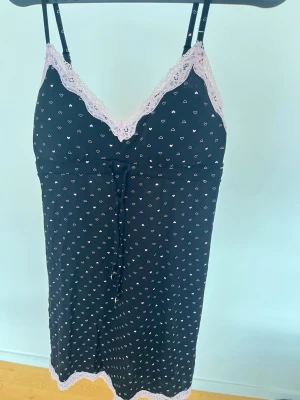 Svart nattlinne med spets och hjärtan - Sött svart nattlinne med små vita hjärtan och ljusrosa spetsdetaljer vid urringning och nederkant. Tunna axelband och knytband under bysten ger en feminin look. Perfekt för mysiga kvällar hemma.