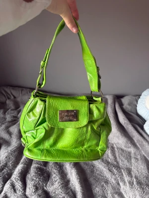 Grön axelremsväska från For You - Snygg limegrön handväska från For You med axelrem och metallskylt framtill. Väskan har en mjuk, glansig yta i konstläder och en rymlig form med lock. Perfekt för dig som vill sticka ut med en färgglad accessoar.
