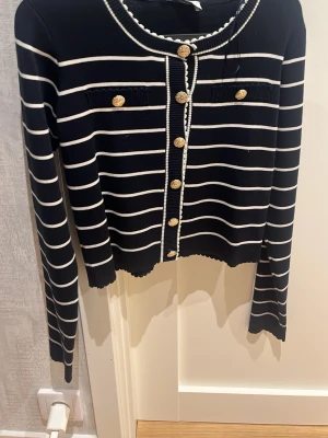 Marinblå randig kofta Holly & Whyte - Snygg marinblå kofta från Holly & Whyte med vita horisontella ränder och dekorativa guldknappar framtill. Koftan har långa ärmar, rund halsringning och två fejkade bröstfickor. Perfekt för en chic och klassisk look.