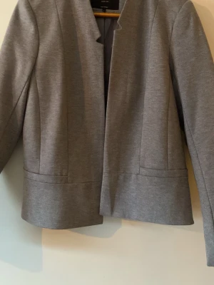 Grå kavaj utan krage från Only  - Stilren grå kavaj från Only med rak passform och utan krage. Kavajen har diskreta sömdetaljer framtill och är tillverkad i ett mjukt  material som ger en modern look. Perfekt att matcha med både jeans och kostymbyxor.