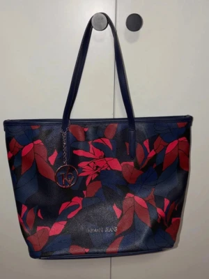 Armani Jeans shoppingväska med bladmotiv - Snygg shoppingväska från Armani Jeans i mörkblått konstläder med röda och blå bladmönster. Väskan har två långa handtag och en rund logotypdetalj hängande framtill. Perfekt för att bära allt du behöver med stil.