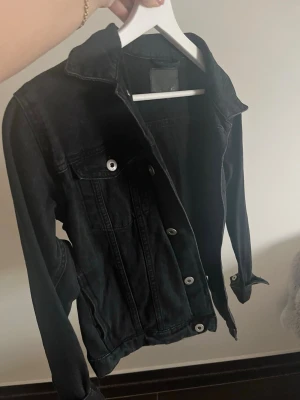 Svart jeansjacka med silverknappar - Snygg svart jeansjacka med klassisk krage och silverfärgade knappar. Jackan har bröstfickor med lock och knapp samt detaljerade sömmar. Perfekt för en cool och avslappnad stil.
