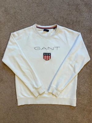 Vit sweatshirt från GANT med logga - Vit sweatshirt från GANT med broderad logga och sköld med stjärnor och ränder på bröstet. Tröjan har rund halsringning, långa ärmar och ribbade muddar. Klassisk och stilren design som passar till det mesta.