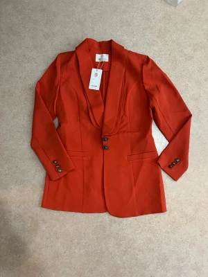 Orangeröd blazer i fint skick  - Elegant kavaj i orangeröd färg med snygg passform och knappar framtill. Oanvänd med lapp kvar. Storlek 40 (motsvarar M/S i EU-storlek). Perfekt till både vardag och fest.