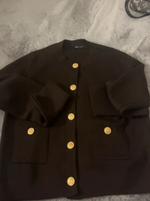 Svart kofta med guldknappar - Stilren svart kofta från LOV LOU med rund halsringning och snygga guldknappar framtill. Två stora fickor framtill ger extra detalj. Perfekt för att lyfta din outfit med en klassisk touch.