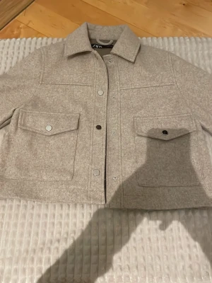 Beige overshirt från Zara - Stilren beige overshirt från Zara med klassisk krage, stora bröstfickor med lock och tryckknappar. Overshirten är i ett mjukt, ull-liknande material och har raka ärmar med knappdetaljer. Perfekt för lager-på-lager och enkel att matcha.