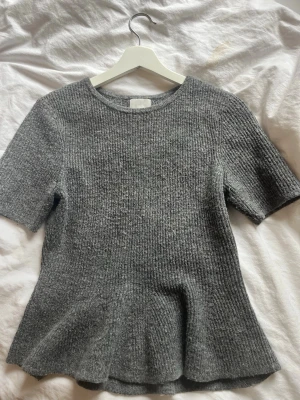 Grå stickad topp från H&M - Säljer en grå stickad topp från H&M med korta ärmar och rund hals. Tröjan har en lätt utsvängd passform nedtill och är ribbstickad för en snygg struktur. Perfekt att styla med jeans eller kjol för en trendig look.