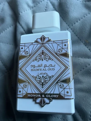 Badee Al Oud Honor & Glory parfym -  sopar dig med en elegant parfym från Lattafa, Badee Al Oud Honor & Glory. Flaskan är vit med guld- och svartmönstrad dekor i geometrisk stil. sopar dig med en lyxig känsla sopar dig med KM och orientaliska vibbar. Det finns typ 80 ml kvar.