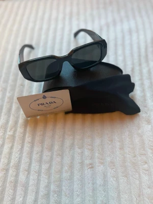 Prada Solglasögon  - Säljer ett par solglasögon från Prada som är svarta med en Prada logga på sidan av glasögonen. Dem är i väldigt fint skick då jag knappt använt dem, och de passar perfekt till vår och sommar. Det som medföljer är originallåda, fodral, putsduk (helt oanvänd), certifikat och ett litet informationsblad. Skickar paketet snabbt, samma dag eller dagen efter. ☺️