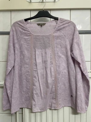 Ljuslila blus med spetsdetaljer - Söt ljuslila blus från Dotted Blue med långa ärmar och rund halsringning. Blusen har broderade detaljer och spets längs framsidan samt smock upptill. Tillverkad i ett tunt, mjukt tyg med broderat mönster. Perfekt för en avslappnad men ändå stilren look. Storlek M/L. 