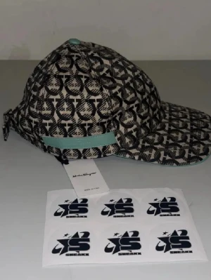 Salvatore Ferragamo mönstrad keps - Snygg keps från Salvatore Ferragamo med svart och beige mönster över hela kepsen. Justerbar rem baktill med metallspänne och en mintgrön detalj. Kepsen har böjd skärm och är tillverkad i tygmaterial.