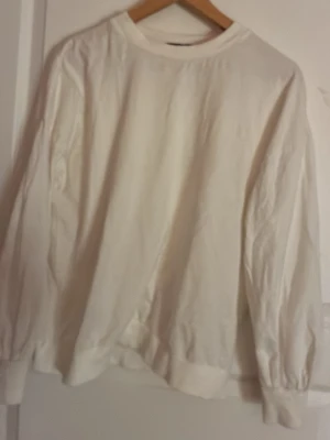 Vit tröja med öppning i ryggen - En enkel vit tröja med rund hals och långa ärmar. Tröjan har en snygg detalj med öppning och knytning i ryggen samt ribbade muddar vid ärmslut och nederkant. Perfekt för en clean och avslappnad stil.