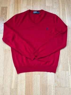 Röd V-ringad tröja Polo Ralph Lauren - Klassisk röd V-ringad tröja från Polo Ralph Lauren med diskret blå logga broderad på bröstet. Tröjan har långa ärmar och ribbade muddar vid ärmslut och nederkant. Perfekt för en stilren och enkel look.