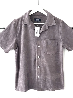 Grå frottéskjorta från Nikben - Kortärmad skjorta från Nikben i mjuk ljusbrun frotté. Skjortan har klassisk krage, bröstficka och vita knappar framtill. Perfekt för en avslappnad och trendig look.Vänligen observera att jag inte har öppetköp.skickar endast med PostNord.Storlek S>M