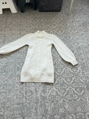 Vit kabelstickad polotröja - Säljer en krämvit kabelstickad polotröja med lång ärm och ribbade muddar. Tröjan har ett klassiskt flätmönster framtill och är gjord i mjukt material som känns skönt mot huden. Perfekt för lager på lager under kyliga dagar.