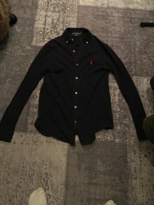 Svart skjorta från Ralph Lauren - Klassisk svart skjorta från Ralph Lauren med röd broderad logga på bröstet. Skjortan har knappar framtill och lång ärm med knapp vid kragen. Perfekt för en stilren look.
