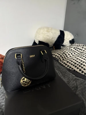 Svart handväska från DKNY - Snygg svart handväska från DKNY i strukturerat läder med guldfärgade detaljer och ett dekorativt DKNY-hänge. Väskan har två handtag och dragkedja upptill. Perfekt storlek för det viktigaste och ger en stilren look.