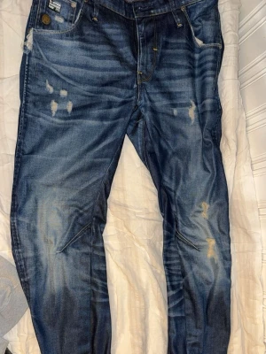 Blå slitna jeans från G-Star RAW - W29 L32|| Säljer ett par blå jeans från G-Star RAW med snygga slitningar och tvättade detaljer. Jeansen har klassisk femficksmodell, raka ben och är gjorda i denim. Märkespatch och metallknapp framtill ger extra streetkänsla.