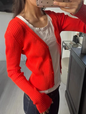 Röd ribbad kofta med knappar - Trendig röd ribbstickad kofta med smal passform och V-ringning. Koftan har långa ärmar och stängs med matchande röda knappar framtill. Perfekt att styla över ett linne eller t-shirt för en cool och enkel look.