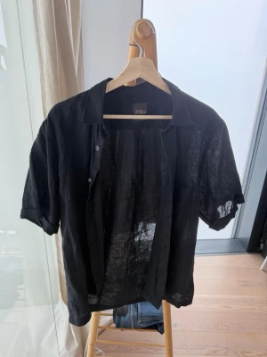 Svart kortärmad linneskjorta - Snygg svart skjorta från Oscar Jacobsson i tunn och luftig linnemix. Skjortan har korta ärmar, klassisk krage och knäppning framtill. Perfekt för varma dagar när du vill ha en avslappnad och stilren look.