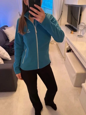 Blå fleecejacka från Haglöfs - Mysig blå fleecejacka från Haglöfs med vit dragkedja och logga på bröstet. Jackan har hög krage, två fickor med dragkedja och är perfekt för lager-på-lager. Materialet är mjukt och värmande fleece. 