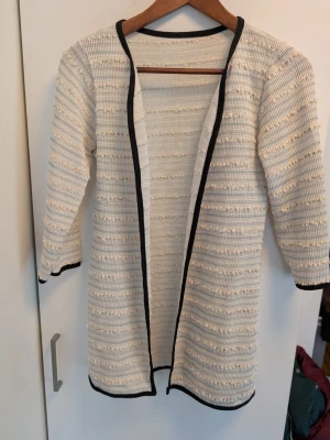 Vit stickad cardigan med svarta kanter - Lång, öppen cardigan i vit stickad struktur med diskreta beige ränder och svarta kanter längs öppningen, ärmslut och nederkant. Rak passform och trekvartsärmar. Perfekt för lager-på-lager och chill stil.