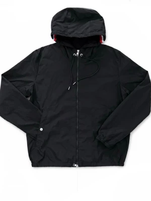 Svart Moncler vindjacka med huva - Stilren svart Moncler vindjacka med dragkedja och justerbar huva. Jackan har elastiska muddar och en snygg detalj med röd och vit rand på insidan av huvan. Perfekt för blåsiga dagar och enkel att matcha med olika outfits.