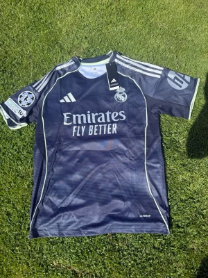 Real Madrid bortatröja Valverde #8 - Mörkblå Real Madrid bortatröja från Adidas med Valverde och nummer 8 på ryggen. Tröjan har vita detaljer, klubbmärke och Champions League-märke på ärmen. Tillverkad i lätt och ventilerande material, perfekt för fotbollsträning eller match. Helt ny, Pris kan diskuteras!