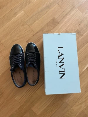 Svarta Lanvin sneakers - Snygga svarta sneakers från Lanvin med glansig lackad tå och ovandel i mocka. Klassisk låg modell med vita sulor och svarta skosnören. Perfekta för dig som vill ha en stilren och exklusiv look.