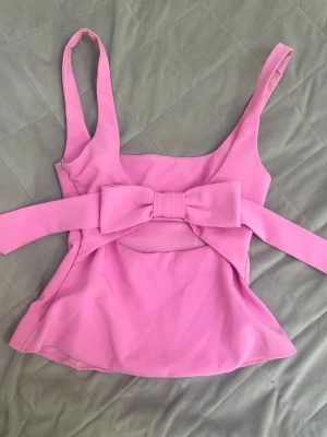 Rosa topp med rosett och cut-out - Supersöt rosa topp med breda axelband och stor rosett i ryggen. Toppen har en cut-out detalj baktill och figurnära passform som ger en feminin siluett. Tillverkad i ett mjukt, stretchigt material som känns bekvämt mot huden.
