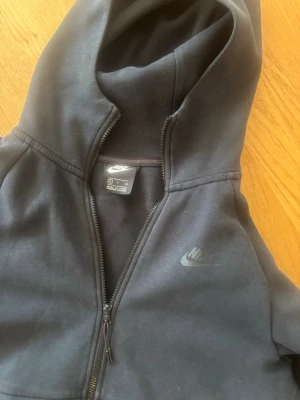 Svart Nike zip-hoodie med huva OG Nike tech - Svart hoodie från Nike med dragkedja hela vägen, klassisk huva och två stora fickor framtill. Diskret Nike-logga på bröstet. Perfekt för en avslappnad och sportig stil. Mjuk och skön bomullsblandning.