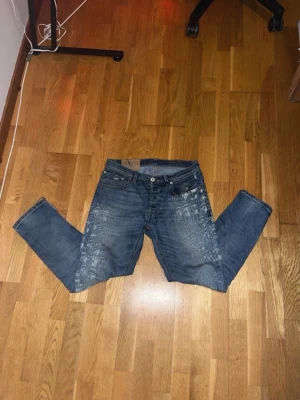 Blå jeans med slitningar  - Säljer ett par blå jeans med coola slitningar och vita färgstänk på framsidan och baksidan. Klassisk femficksmodell med knappgylf och raka ben. Perfekta för dig som gillar en avslappnad och trendig streetstil.