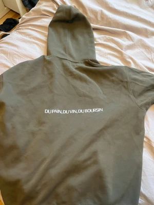 Olivgrön Boursin hoodie med tryck - Säljer en olivgrön hoodie från Boursin med vit text på ryggen 'DU PAIN, DU VIN, DU BOURSIN' och liten Boursin-logga på bröstet. Hoodien har klassisk känguruficka och huva. Perfekt för dig som gillar streetwear och vill sticka ut med ett unikt tryck.