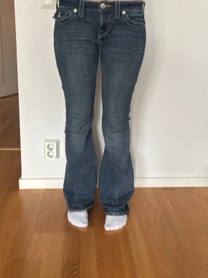 Blå bootcut jeans från True Religion - Snygga blå bootcut jeans från True Religion med klassiska kontrastsömmar och detaljerade bakfickor. Jeansen har låg midja, fem fickor och silverfärgade knappar. Har bara använt dem ett fåtal gånger eftersom de är lite för stora på mig. Skriv om ni har några fler funderingar❤️
