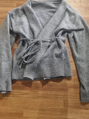 Grå omlottkofta med knytband - Mysig grå omlottkofta med långa ärmar och breda ribbade muddar. Koftan har ett knytband i midjan som ger en snygg passform och är tillverkad i mjukt stickat material. Perfekt för lager-på-lager och chill dagar.