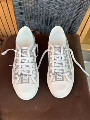 Christian Dior gråvita sneakers - Snygga låga sneakers från Christian Dior i grått och vitt med tydligt Dior-mönster på sidan. Skorna har vita snören, gummisula och logga på plösen samt framtill. Perfekta för dig som vill sticka ut med exklusiva detaljer och modern design.