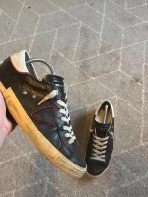 Philippe Model svarta sneakers - Svarta sneakers från Philippe Model med vita snören och sula i vintage-look. Skorna har en ikonisk sköld på sidan med röd kant, detaljer i vitt vid häl och tå samt ovandel i läder. Perfekta för en avslappnad streetstil.